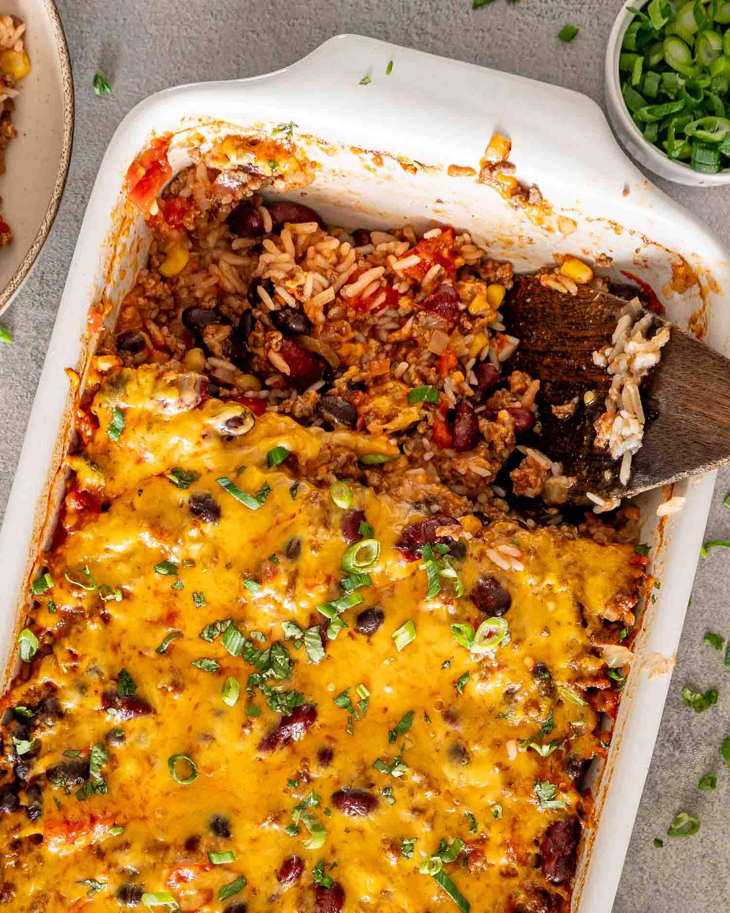 Overhead view of partially served chili rice bake with melted cheddar, green onions, and a wooden spoon inside the dish.