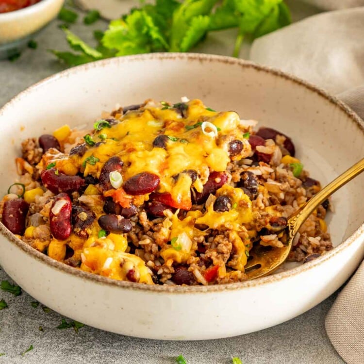 Chili rice bake served in a cream ceramic bowl with melted cheddar, beans, corn, herbs, and a gold fork.
