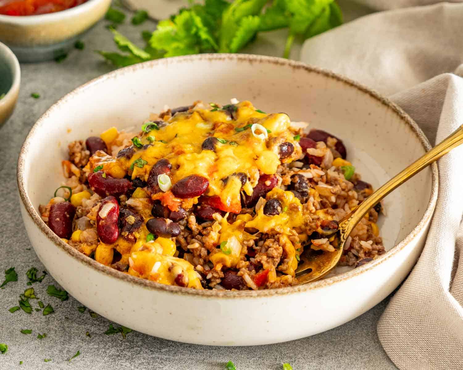Chili rice bake served in a cream ceramic bowl with melted cheddar, beans, corn, herbs, and a gold fork.