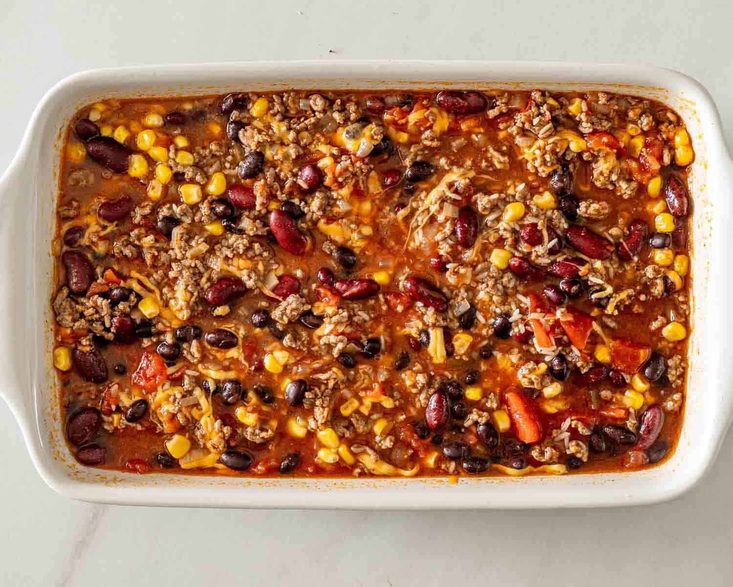 Baked chili rice mixture with ground beef, kidney beans, black beans, corn, tomatoes, and broth in a white dish.