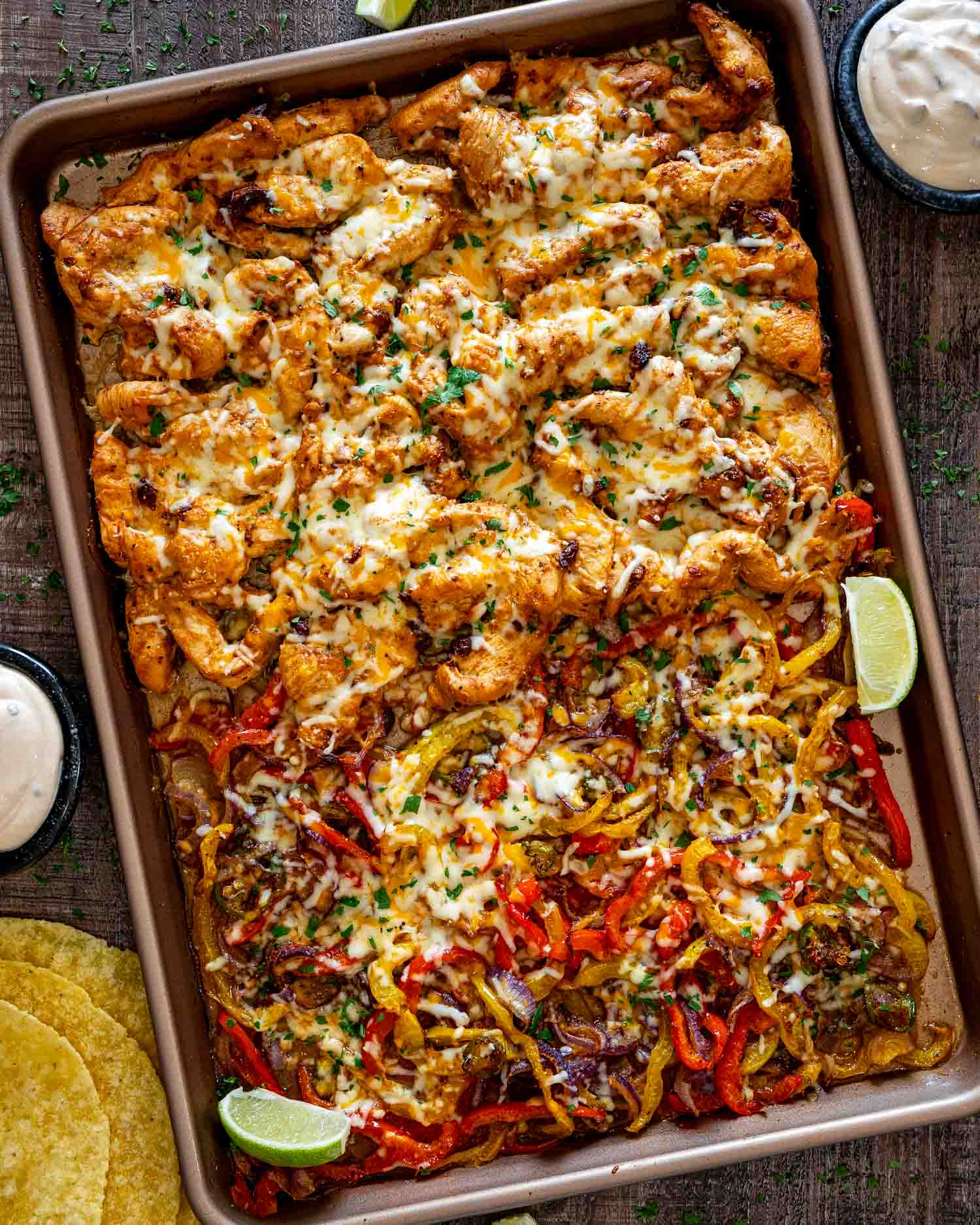 Overhead view of sheet pan chipotle chicken with melted cheese, colorful roasted peppers, red onions, cilantro, and lime wedges.