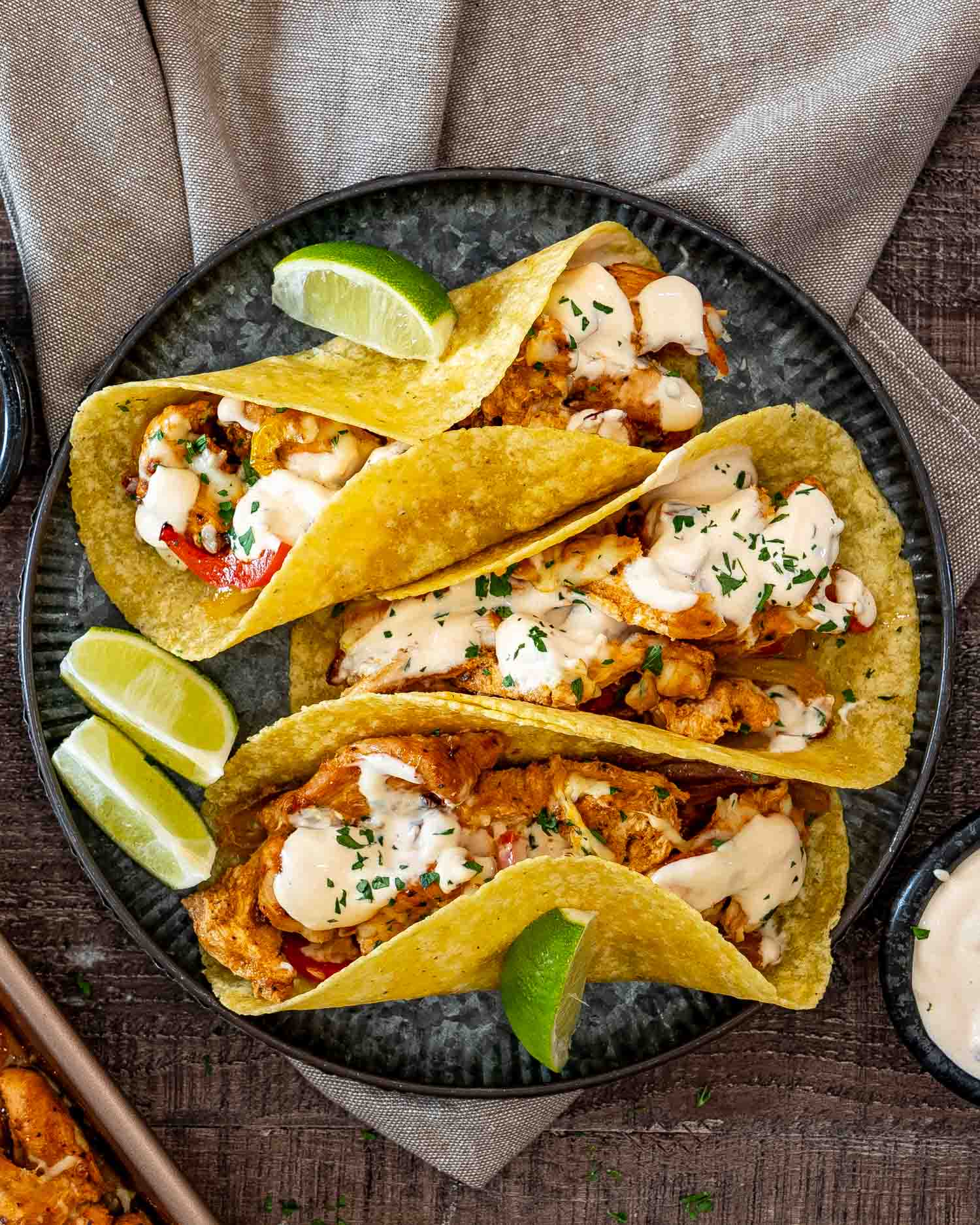 Three chipotle chicken tacos on a dark plate, drizzled with creamy sauce and garnished with cilantro and lime wedges.