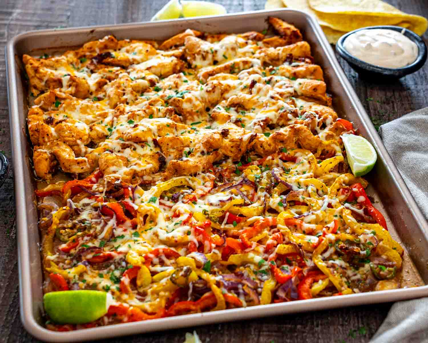 Sheet pan chipotle chicken with melted cheese, roasted bell peppers, red onions, and cilantro, served alongside creamy sauce.