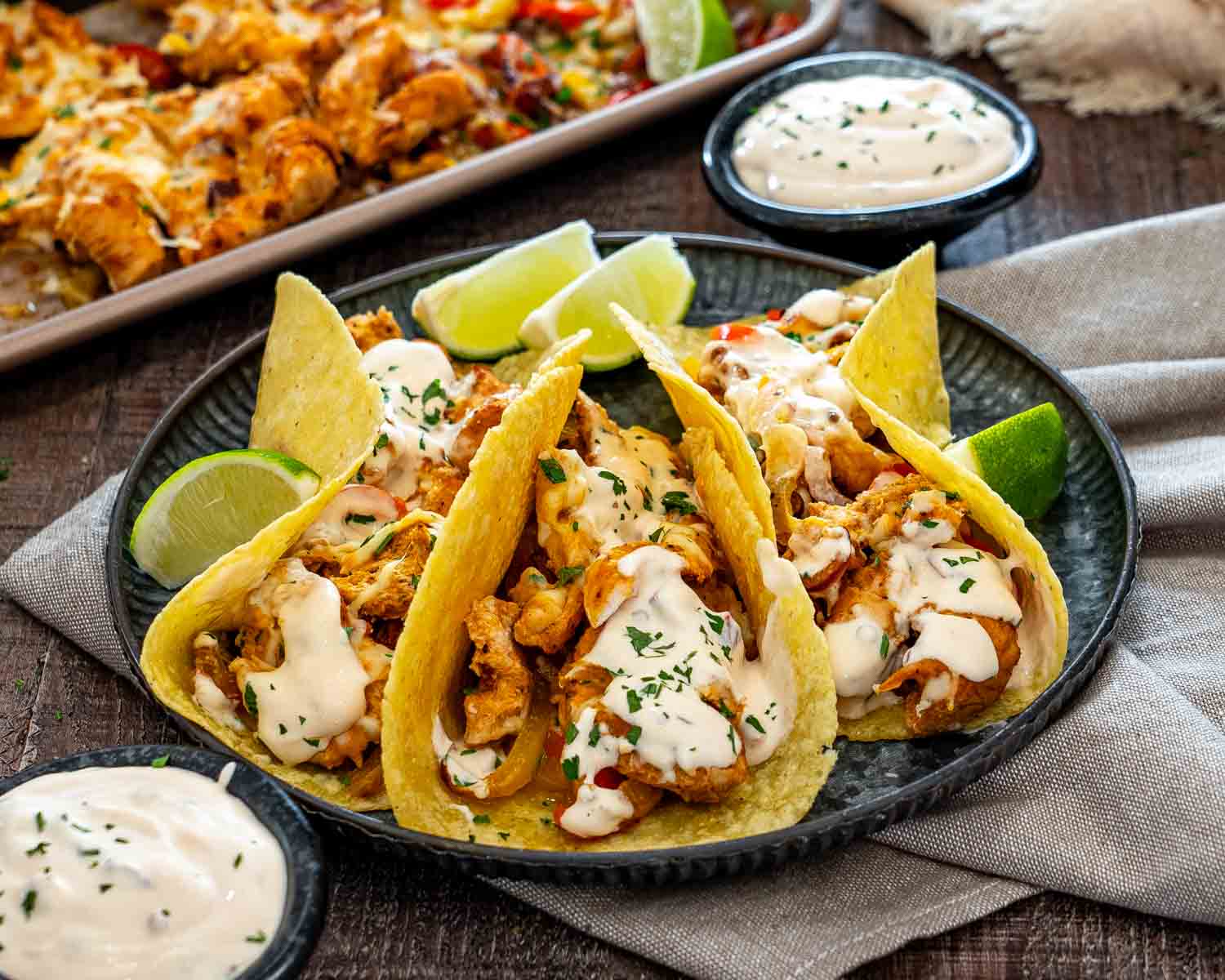 Chipotle chicken tacos topped with creamy sauce and cilantro, served with lime wedges beside a sheet pan of filling.
